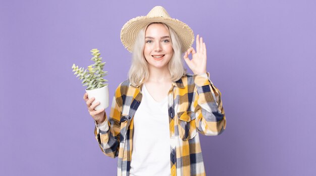 Молодая красивая женщина-альбинос чувствует себя счастливой, демонстрирует одобрение жестом и держит кактус из комнатного растения