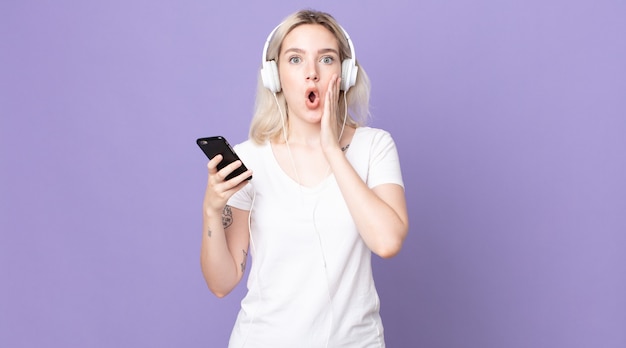 Young pretty albino woman feeling shocked and scared with headphones and smartphone