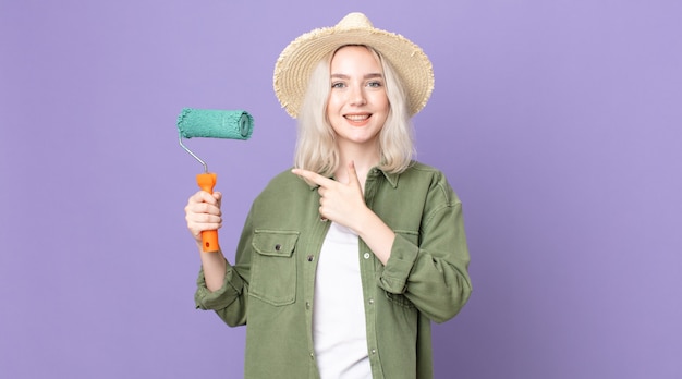 Young pretty albino woman looking excited and surprised pointing to the side and holding a roller paint