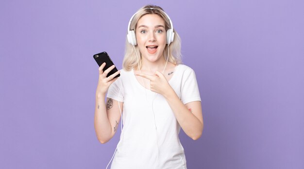 Young pretty albino woman looking excited and surprised pointing to the side with headphones and smartphone