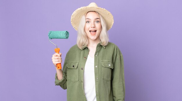 Young pretty albino woman looking happy and pleasantly surprised and holding a roller paint