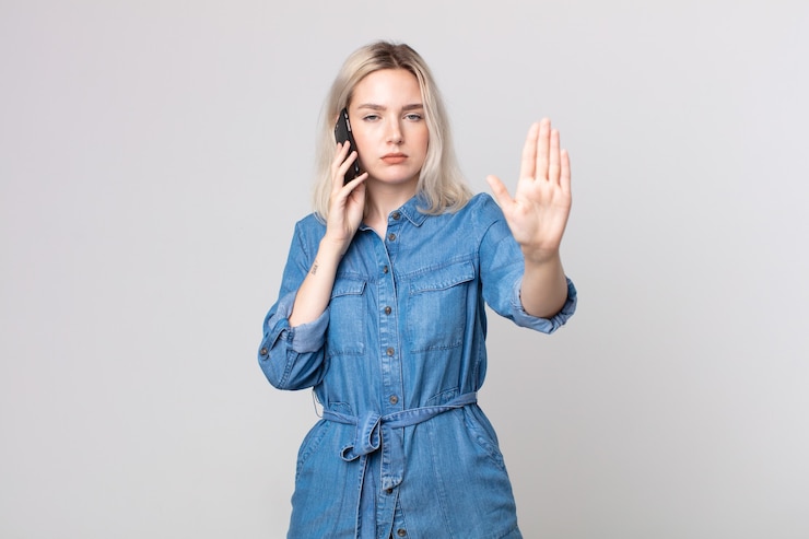 Photo young pretty albino woman looking serious showing open palm making stop gesture and talking with a smartphone