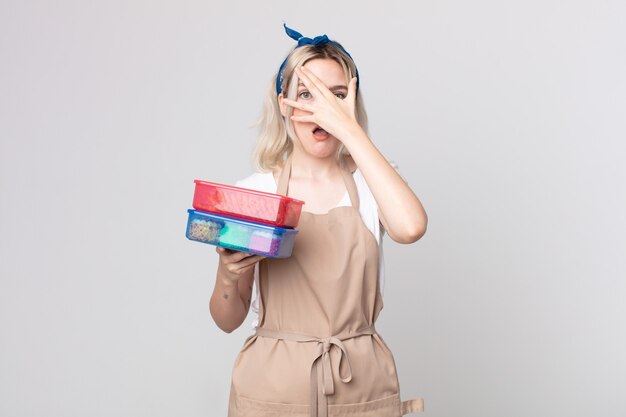 Young pretty albino woman looking shocked, scared or terrified, covering face with hand holding food tupperwares