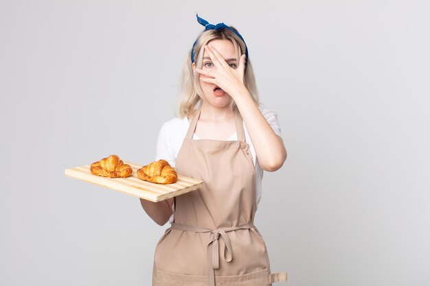 Young pretty albino woman looking shocked, scared or terrified, covering face with hand with a croissants tray