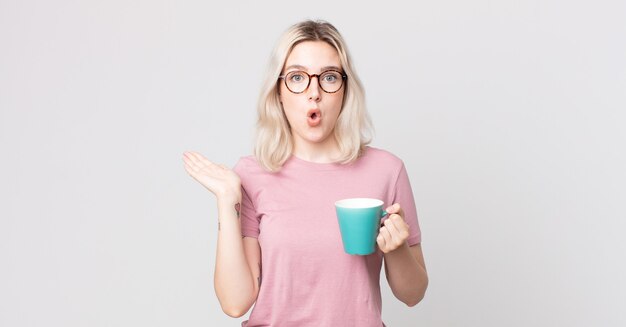 Young pretty albino woman looking surprised and shocked, with jaw dropped holding an object with a coffee mug