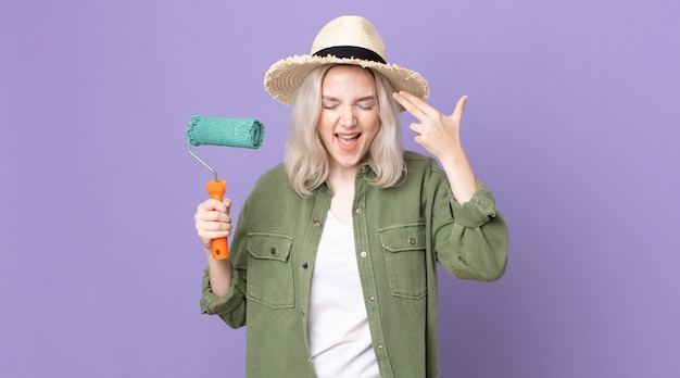 Young pretty albino woman looking unhappy and stressed, suicide gesture making gun sign and holding a roller paint