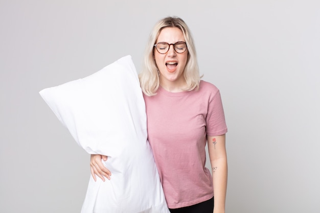 Young pretty albino woman shouting aggressively, looking very angry wearing pajamas and holding a pillow