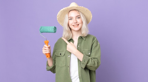 Young pretty albino woman smiling cheerfully, feeling happy and pointing to the side and holding a roller paint