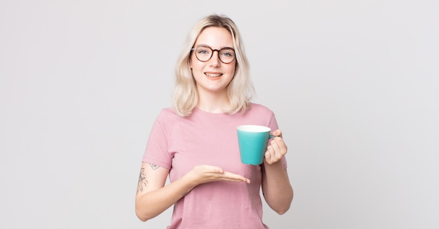 Giovane bella donna albina che sorride allegramente, si sente felice e mostra un concetto con una tazza da caffè