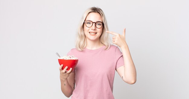 Young pretty albino woman smiling confidently pointing to own broad smile with a breakfast bowl