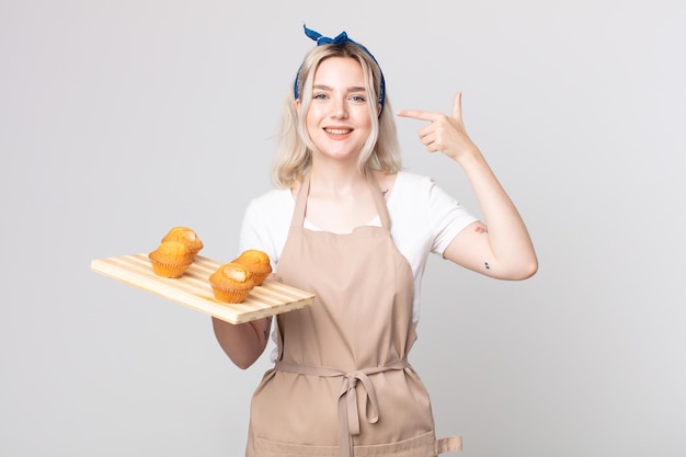 Young pretty albino woman smiling confidently pointing to own broad smile with a muffins tray