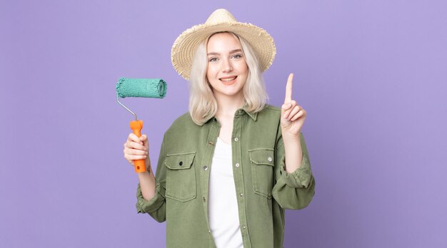 Young pretty albino woman smiling and looking friendly, showing number one and holding a roller paint