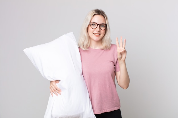 Young pretty albino woman smiling and looking friendly, showing number three wearing pajamas and holding a pillow