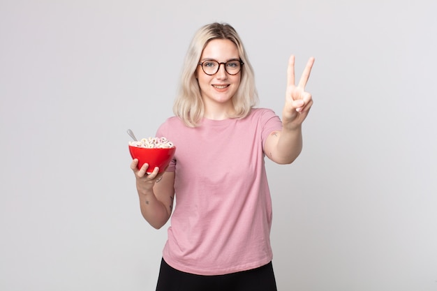 Young pretty albino woman smiling and looking happy, gesturing victory or peace with a breakfast bowl