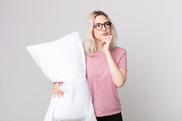 Young pretty albino woman thinking, feeling doubtful and confused wearing pajamas and holding a pillow