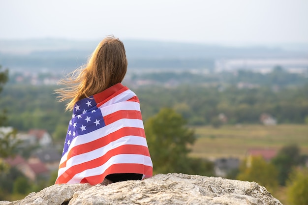 Giovane bella donna americana con i capelli lunghi che tengono sventolando la bandiera degli stati uniti del vento sui suoi sholders che riposano all'aperto godendosi la calda giornata estiva.