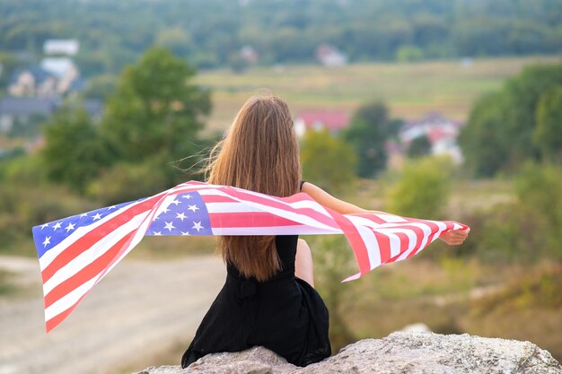 Photo young pretty american woman with long hair holding waving on wind usa flag on her sholders resting outdoors enjoying warm summer day