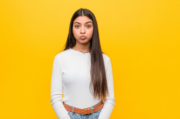 Young pretty arab woman against a yellow wall blows cheeks, has tired expression. Facial expression concept.