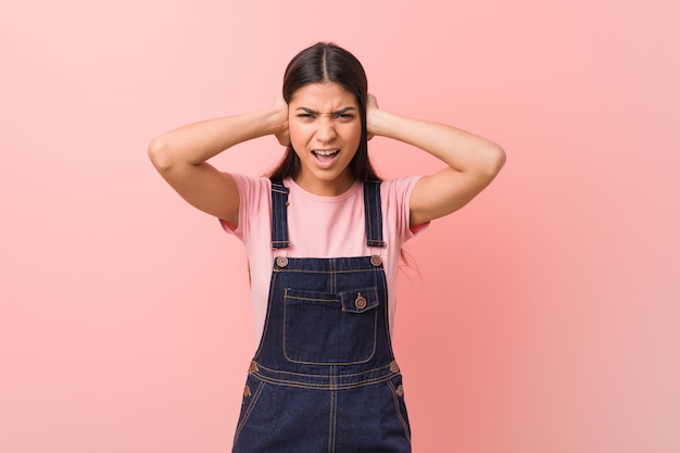 Young pretty arab woman wearing a jeans dungaree covering ears with hands trying not to hear too loud sound.