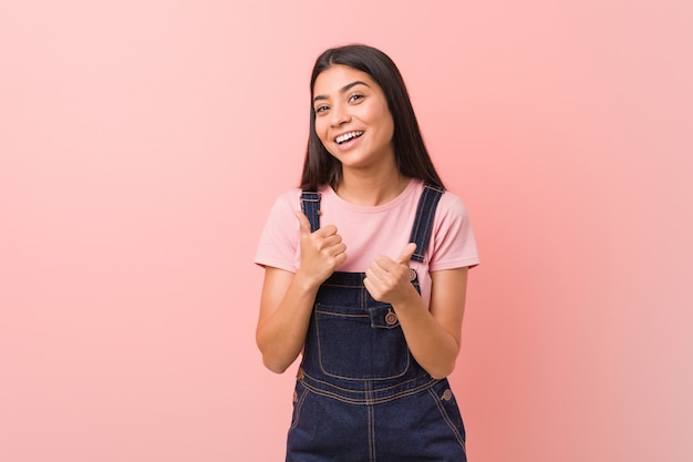 Young pretty arab woman wearing a jeans dungaree raising both thumbs up, smiling and confident.