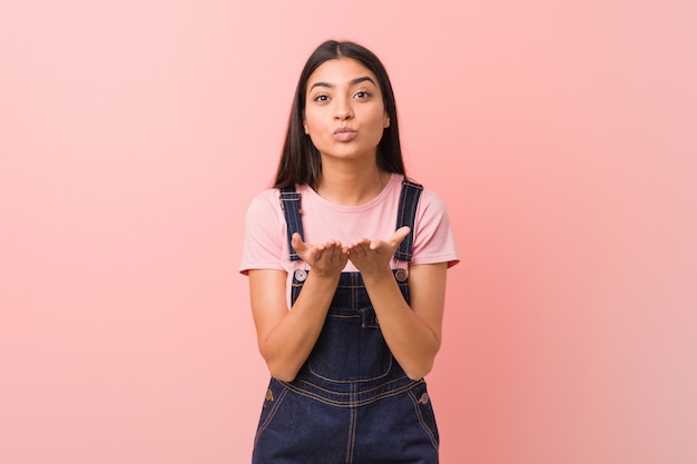 Young pretty arab woman wearingjeans dungaree folding lips and holding palmssend air kiss.