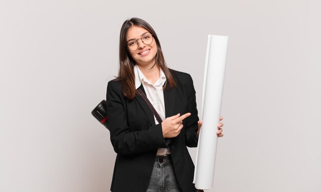 young pretty architect woman smiling cheerfully, feeling happy and pointing to the side and upwards, showing object in copy space