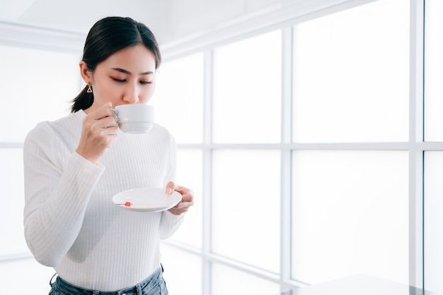 A young pretty Asian female wearing a sweater drinking a cup of hot coffee.