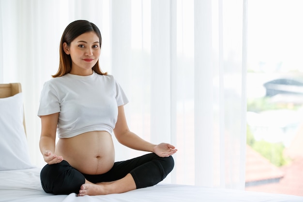 Giovane bella donna incinta asiatica seduta su un letto facendo esercizio deliziosamente dallo yoga. sta sorridendo e guardando una macchina fotografica. concetto di madre felice sana.