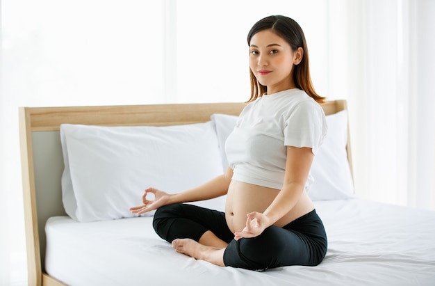 Young pretty Asian pregnant woman sitting on a bed doing exercise delightfully by Yoga. She is smiling and looking at a camera. Healthy happy mother concept.