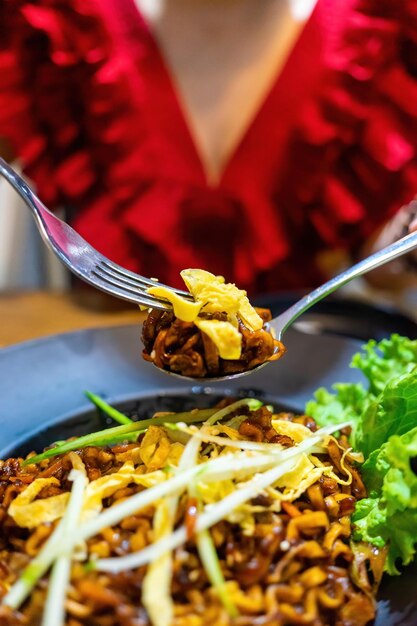 Young pretty asian woman hand holds and eats dry dark soy\
noodles (local name is zajang noodle) with shredded egg crepes in\
restaurant in vietnam. food and drink concept.