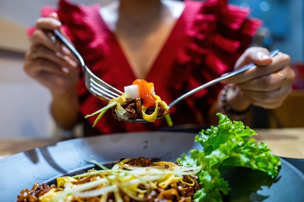 Young pretty asian woman hand holds and eats dry dark soy\
noodles (local name is zajang noodle) with shredded egg crepes in\
restaurant in vietnam. food and drink concept.