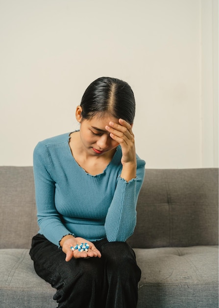 Young Pretty Asian woman holding medicine pills when sitting on couch feeling headache