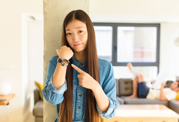 Young pretty asian woman looking impatient and angry, pointing at watch, asking for punctuality, wants to be on time