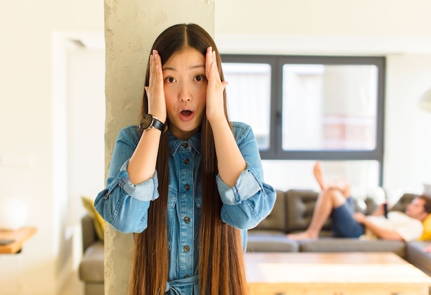 불쾌하게 충격을 받거나 무서워하거나 걱정하고 입을 벌리고 손으로 두 귀를 덮고있는 젊은 꽤 아시아 여자