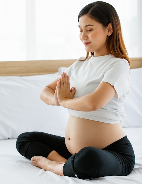 ベッドに座っている若いかなりアジアの女性は、カメラを見ている笑顔で彼女の胸の前で彼女の手を結合します。ヨガは妊娠中の女性にとって素晴らしい運動です。