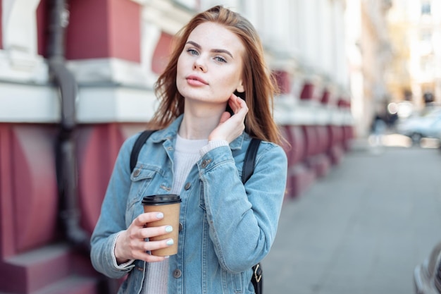 市内でコーヒーカップを保持しているデニムジャケットを着た若いかなり魅力的な女性