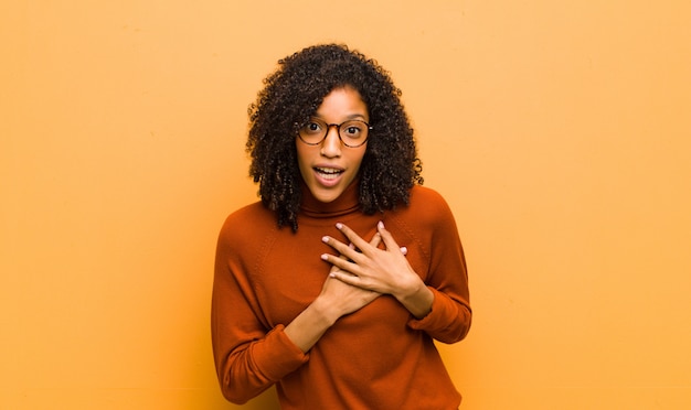 Young pretty black woman feeling shocked and surprised, smiling, taking hand to heart, happy to be the one or showing gratitude against orange wall