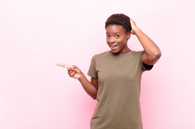 Young pretty black woman laughing pointing to lateral blank space against pink wall