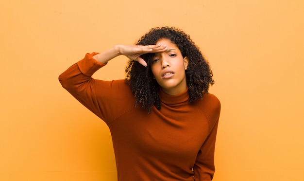 Young pretty black woman looking bewildered and astonished, with hand over forehead looking far away, watching or searching against orange wall