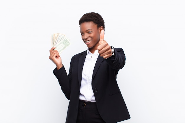 Young pretty black womanfeeling proud, carefree, confident and happy, smiling positively with thumbs up with dollar banknotes