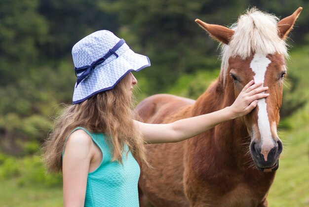 ぼやけた緑の日当たりの良い夏に美しい栗の馬を愛sunする太陽帽子の若いかなりブロンドの長髪の女性。動物、ケア、友情、誠実さと農業の概念が大好きです。