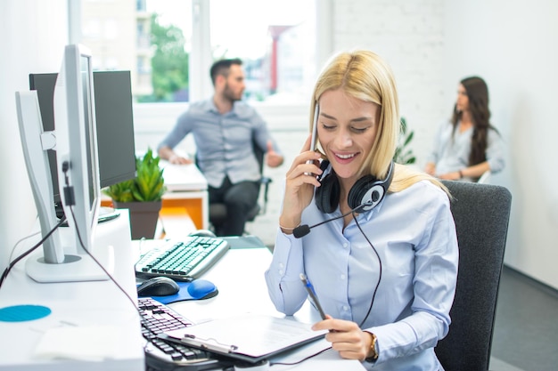 young pretty blond woman sitting at office desk and reading important paper on clipboard while talking on mobile phone