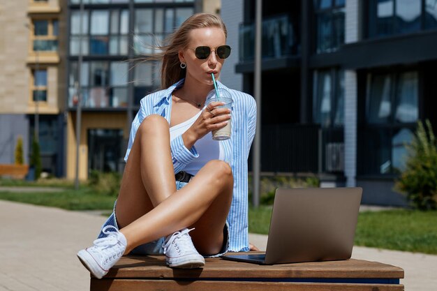 Фото Молодая симпатичная блондинка в солнечных очках решает бизнес-задачи с помощью ноутбука на улице