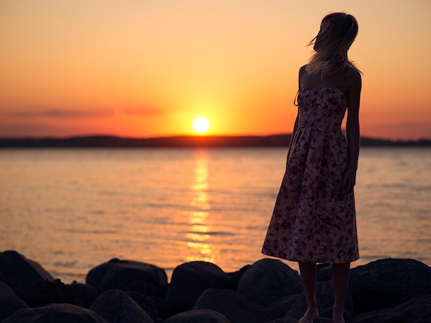 Photo young and pretty blonde girl standing on the shore in a dress and admiring the sunset