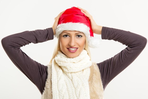 Photo a young pretty blonde girl with a santa's cap smiling looking at the camera while her hands puts a cap on her head.