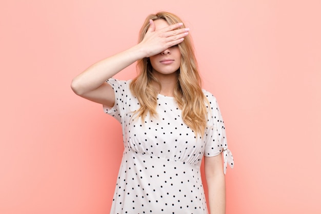 Young pretty blonde woman covering eyes with one hand feeling scared or anxious, wondering or blindly waiting for a surprise against flat color wall