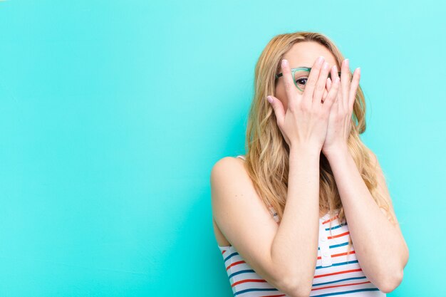 Young pretty blonde woman covering face with hands, peeking between fingers with surprised expression and looking to the side against flat color wall