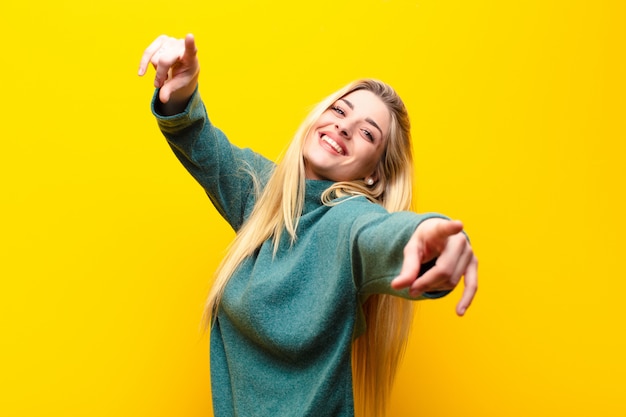 Photo young pretty blonde woman feeling happy and confident, pointing to camera with both hands and laughing, choosing you over yellow wall