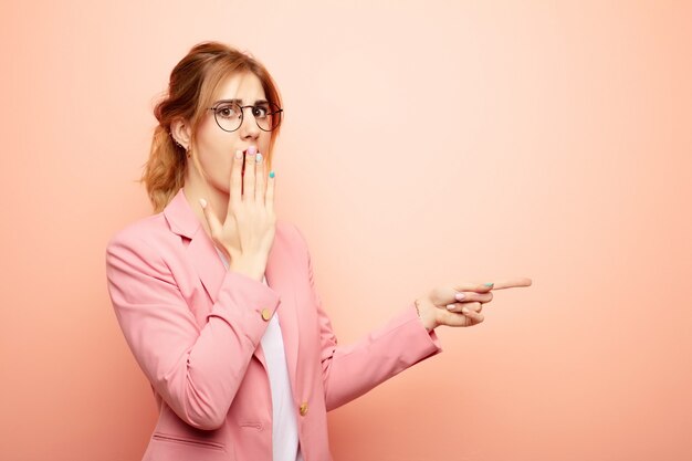 Young pretty blonde woman feeling happy, shocked and surprised, covering mouth with hand and pointing to lateral copy space. business concept