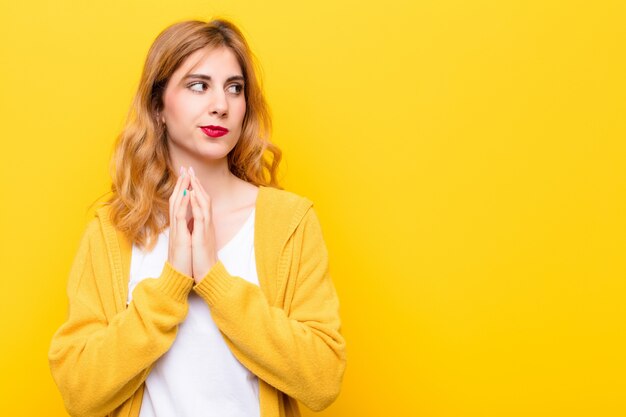 Young pretty blonde woman feeling proud, mischievous and arrogant while scheming an evil plan or thinking of a trick over yellow wall